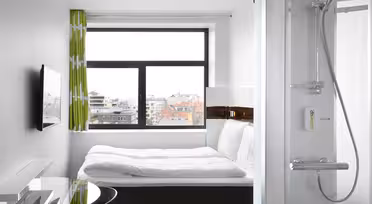Kleines, modernes Hotelzimmer mit großem Fenster und Blick auf die Stadt. Das Zimmer verfügt über ein ordentlich gemachtes Bett, einen an der Wand montierten Fernseher und einen Glasschreibtisch. Im Vordergrund ist eine kompakte Dusche zu sehen. Grüne Vorhänge sorgen für einen Hauch von Farbe.