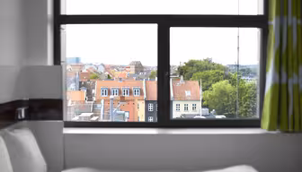 Ein Blick durch ein Fenster auf eine Stadtlandschaft mit Gebäuden mit roten Dächern und üppigen Bäumen. Im Vordergrund ist ein Bett mit weißer Bettwäsche und teilweise grün-weißen Vorhängen zu sehen.