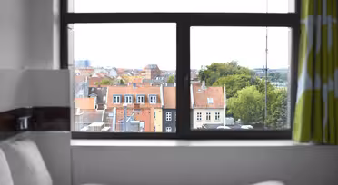 Ein Blick durch ein Fenster auf eine Stadtlandschaft mit Gebäuden mit roten Dächern und üppigen Bäumen. Im Vordergrund ist ein Bett mit weißer Bettwäsche und teilweise grün-weißen Vorhängen zu sehen.