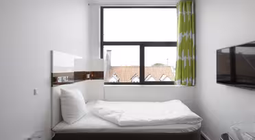 Ein modernes, minimalistisches Schlafzimmer mit einem Einzelbett vor einem Fenster. Das Bett ist mit weißer Bettwäsche und einem Kissen ausgestattet. Es gibt einen grünen Vorhang mit weißem Muster, einen an der Wand montierten Flachbildfernseher und einen kleinen Beistelltisch aus Glas.
