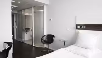 Ein modernes Hotelzimmer verfügt über ein ordentlich gemachtes Bett mit weißer Bettwäsche, einen eleganten schwarzen Stuhl und einen runden Glastisch. Im Hintergrund befindet sich ein verglastes Badezimmer mit Dusche und Wandarmaturen.