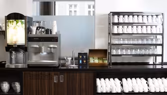 Eine moderne Kaffeestation mit zwei Maschinen auf einer Theke aus dunklem Holz, umgeben von Stapeln weißer Tassen und Gläser auf Regalen. Zwischen den Maschinen stehen eine Schachtel Tee und ein Behälter mit Rührstäbchen.