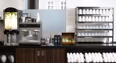 Eine moderne Kaffeestation mit zwei Maschinen auf einer Theke aus dunklem Holz, umgeben von Stapeln weißer Tassen und Gläser auf Regalen. Zwischen den Maschinen stehen eine Schachtel Tee und ein Behälter mit Rührstäbchen.