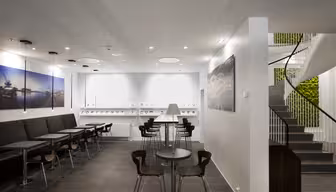 Modernes Café-Interieur mit schwarzen Sitzgelegenheiten, eleganten Tischen und Stühlen mit Holzakzenten. Eine Wendeltreppe führt hinauf, und ein vertikaler Garten ist sichtbar. Große Fotografien von Stadtlandschaften schmücken die Wände. Die moderne Beleuchtung schafft eine helle Atmosphäre.