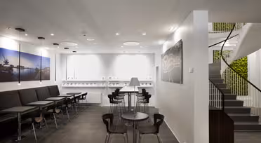 Modernes Café-Interieur mit schwarzen Sitzgelegenheiten, eleganten Tischen und Stühlen mit Holzakzenten. Eine Wendeltreppe führt hinauf, und ein vertikaler Garten ist sichtbar. Große Fotografien von Stadtlandschaften schmücken die Wände. Die moderne Beleuchtung schafft eine helle Atmosphäre.