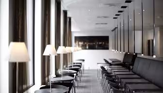Ein modernes, minimalistisches Café mit Reihen kleiner Tische und Stühle. Jeder Tisch hat eine kleine Lampe. Große Fenster entlang der linken Seite, während auf der rechten Seite eine lange Bank verläuft. Der Raum ist gut beleuchtet und elegant gestaltet.