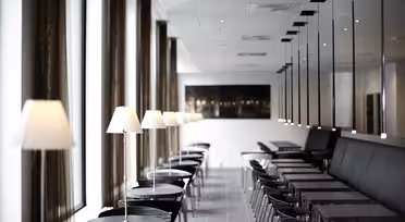 Ein modernes, minimalistisches Café mit Reihen kleiner Tische und Stühle. Jeder Tisch hat eine kleine Lampe. Große Fenster entlang der linken Seite, während auf der rechten Seite eine lange Bank verläuft. Der Raum ist gut beleuchtet und elegant gestaltet.
