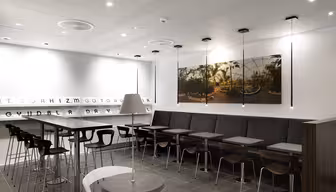 Modernes Café-Interieur mit hohen Tischen und Stühlen, grauem Fliesenboden und weißen Wänden. Ein großes, gerahmtes Naturfoto an der Wand und minimalistische Beleuchtung. Eine lange Sitzbank erstreckt sich entlang einer Raumseite.