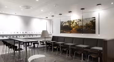 Modernes Café-Interieur mit hohen Tischen und Stühlen, grauem Fliesenboden und weißen Wänden. Ein großes, gerahmtes Naturfoto an der Wand und minimalistische Beleuchtung. Eine lange Sitzbank erstreckt sich entlang einer Raumseite.