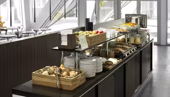 Buffet mit vielfältigen Frühstücksangeboten, darunter Brötchen, Obst, Müsli und Gebäck. Teller und Tassen sind ordentlich gestapelt, ein Toaster ist sichtbar. Das Buffet befindet sich in einem modernen Essbereich mit großen Fenstern und eleganter Einrichtung.