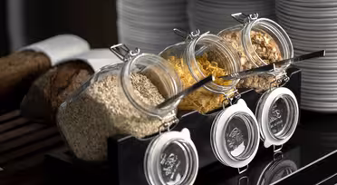 Drei schräg gestellte Gläser mit Haferflocken, Cornflakes und einem gemischten Müsli stehen auf einem schwarzen Ständer. In jedem Glas liegen Löffel. Dahinter sind Stapel weißer Teller und Brotscheiben auf einem Schneidebrett zu sehen.