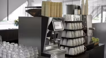 Eine moderne Kaffeemaschine mit Touchscreen ist von Stapeln aus Pappbechern und Keramikbechern umgeben. Klare Gläser sind ordentlich davor angeordnet, und verschiedene Utensilien stehen in der Nähe bereit, was für ein übersichtliches Café-Ambiente sorgt.
