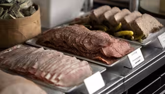 Eine Präsentation von Wurstwaren auf rechteckigen Tellern, darunter Stapel von Salami-, Schinken- und Pastetenscheiben, garniert mit kleinen Essiggurken. Im Hintergrund ist ein Teil eines Behälters mit Gewürzpäckchen zu sehen.