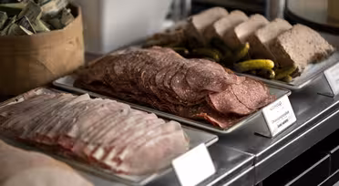 Eine Präsentation von Wurstwaren auf rechteckigen Tellern, darunter Stapel von Salami-, Schinken- und Pastetenscheiben, garniert mit kleinen Essiggurken. Im Hintergrund ist ein Teil eines Behälters mit Gewürzpäckchen zu sehen.