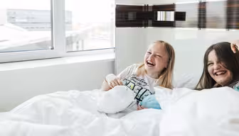 Zwei Mädchen sitzen lachend auf einem Bett neben einem großen Fenster. Sie sind von weißen Decken umgeben und der Raum ist hell und lichtdurchflutet.