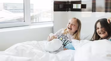 Zwei Mädchen sitzen lachend auf einem Bett neben einem großen Fenster. Sie sind von weißen Decken umgeben und der Raum ist hell und lichtdurchflutet.