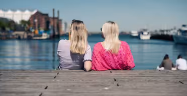 Zwei Personen mit langen blonden Haaren sitzen auf einem Holzsteg und blicken auf ein ruhiges Gewässer mit Booten in der Ferne. Die Person links trägt ein hellviolettes Oberteil, die Person rechts ein leuchtend pinkes. Es ist ein sonniger Tag.