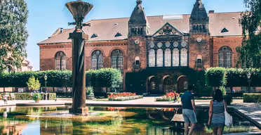 Ein Paar geht auf ein kunstvolles historisches Gebäude mit Bogenfenstern und steilen Dächern zu. Im Vordergrund steht ein großer Brunnen, umgeben von üppigem Grün und Blumen unter einem klaren blauen Himmel.