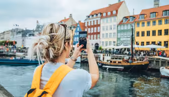 Eine Person mit blonden Haaren und einem gelben Rucksack fotografiert mit ihrem Smartphone farbenfrohe Gebäude entlang der Uferpromenade. Boote liegen im Kanal, Menschen sitzen in Straßencafés. Der Himmel ist bedeckt.