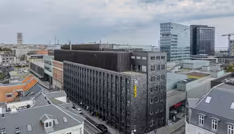 Luftaufnahme einer Stadtlandschaft mit einem modernen, dunkelfarbigen Hotelgebäude mit dem Namen „Wakeup“ an der Seite. Zu den umliegenden Gebäuden gehören ältere Gebäude mit grauen Dächern, moderne Glasgebäude und ein teilweise bewölkter Himmel.
