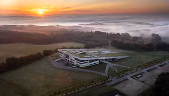 Luftaufnahme eines modernen Gebäudes mit grünem Dach, umgeben von üppigen Feldern und Bäumen. Im Hintergrund geht die Sonne auf und taucht die Landschaft in warmes Licht. Nebel schwebt über den fernen Bäumen und schafft eine ruhige und malerische Szenerie.