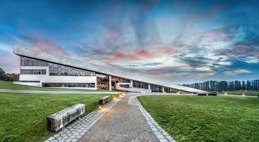 Modernes Gebäude mit schrägem Dach, umgeben von Rasenflächen und sanftem Licht entlang eines Steinwegs. Der Himmel ist mit rosa, blauen und grauen Wolken bemalt und trägt zur ruhigen und modernen Atmosphäre bei.