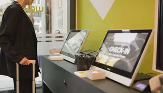 Eine Person checkt an einem Schalter mit zwei Touchscreen-Terminals ein. Sie hält Gepäck in der Hand. Die Terminals zeigen in modernem Ambiente das Wort „CHECK-IN“ vor einer grünen Wand.