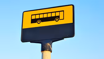 Ein quadratisches gelb-schwarzes Schild mit einem Bussymbol auf einer Stange vor einem klaren blauen Himmel.