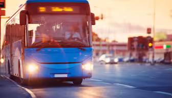 Ein blauer Bus fährt bei Sonnenuntergang durch eine Stadtstraße. Die Bewegung wird durch eine Bewegungsunschärfe eingefangen. Oben auf dem Bus befindet sich ein Zielschild. Im Hintergrund sind im warmen Abendlicht Autos und Gebäude zu sehen.