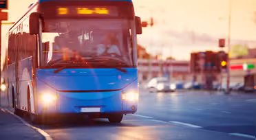 Ein blauer Bus fährt bei Sonnenuntergang durch eine Stadtstraße. Die Bewegung wird durch eine Bewegungsunschärfe eingefangen. Oben auf dem Bus befindet sich ein Zielschild. Im Hintergrund sind im warmen Abendlicht Autos und Gebäude zu sehen.