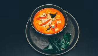 Eine Schüssel leuchtend rotes Curry mit Hähnchenstücken, garniert mit roten Chilischeiben und frischen Kräutern. Die Schüssel steht auf einem passenden dunklen Teller, daneben grüne Blätter, alles vor einem dunklen Hintergrund.