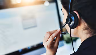 Eine Frau mit Headset sitzt an einem Schreibtisch und konzentriert sich auf einen Computerbildschirm, auf dem Text angezeigt wird. Sie scheint zu sprechen, möglicherweise während eines Telefonats oder einer Videokonferenz, in einem gut beleuchteten Raum.