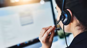 Eine Frau mit Headset sitzt an einem Schreibtisch und konzentriert sich auf einen Computerbildschirm, auf dem Text angezeigt wird. Sie scheint zu sprechen, möglicherweise während eines Telefonats oder einer Videokonferenz, in einem gut beleuchteten Raum.