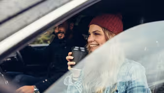 Eine Frau mit einer roten Mütze und einer Kaffeetasse in der Hand sitzt lächelnd auf dem Beifahrersitz eines Autos. Ein Mann in einer schwarzen Jacke fährt. Beide wirken glücklich und sind von der grünen Landschaft vor dem Autofenster umgeben.