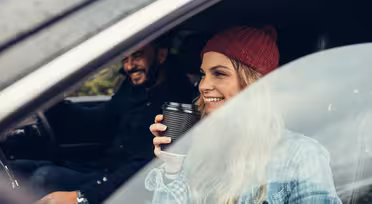 Eine Frau mit einer roten Mütze und einer Kaffeetasse in der Hand sitzt lächelnd auf dem Beifahrersitz eines Autos. Ein Mann in einer schwarzen Jacke fährt. Beide wirken glücklich und sind von der grünen Landschaft vor dem Autofenster umgeben.