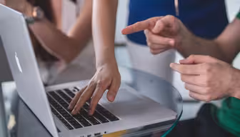 Drei Personen sitzen um einen Glastisch und interagieren mit einem Laptop. Eine Person tippt, während eine andere auf den Bildschirm zeigt und so eine Zusammenarbeit oder Diskussion vorschlägt. Der Fokus liegt auf ihren Händen und dem Laptop.