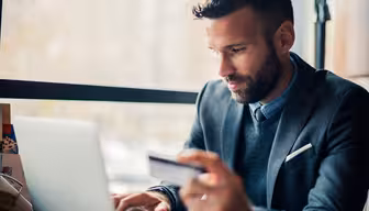 Ein Mann sitzt an einem Tisch am Fenster und konzentriert sich auf seinen Laptop. Er trägt einen grauen Anzug und hält eine Kreditkarte in der Hand, was auf Online-Shopping oder eine Finanztransaktion hindeutet. Der Hintergrund ist leicht verschwommen und unterstreicht eine helle, natürliche Atmosphäre.
