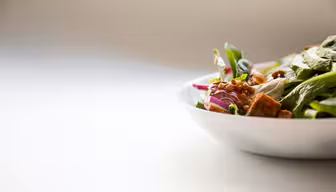 Nahaufnahme einer Salatschüssel vor hellem Hintergrund. Der Salat enthält Spinatblätter, rote Zwiebeln, Croutons, Pilze und Linsen, was ihm ein frisches und farbenfrohes Aussehen verleiht. Die rechte Seite der Schüssel ist sichtbar.