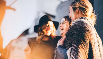 Drei Menschen stehen dicht beieinander und lächeln und lachen im warmen Sonnenlicht. Einer trägt einen Hut, ein anderer eine Brille und der dritte ist in einen Schal gehüllt. Der Hintergrund ist sanft verschwommen und schafft eine fröhliche und freundliche Atmosphäre.