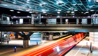 Nachtaufnahme einer modernen, beleuchteten Straßenbahnhaltestelle mit Glasdach. Helle rote und gelbe Lichtstreifen zeigen die Bewegung einer darunter vorbeifahrenden Straßenbahn. Der Bahnsteig ist erhöht und zeigt im Hintergrund einige Personen und einen parkenden Bus.