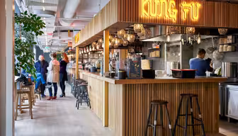 Ein moderner Food Court mit einem Holzstand, auf dem in leuchtenden Buchstaben „Kung Fu“ steht. Der Bereich ist mit hängenden Laternen dekoriert, und an der Theke stehen Hocker. Hinten, in der Nähe anderer Stände, stehen Leute und unterhalten sich.