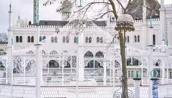 Weiße, kunstvolle Architektur mit Bögen und Kuppeln, teilweise verdeckt von einem kahlen Baum im Vordergrund. Das Bauwerk zeichnet sich durch filigrane Details und einen dekorativen Laternenpfahl vor einem bewölkten Himmel aus.