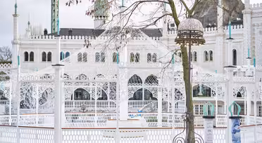 Weiße, kunstvolle Architektur mit Bögen und Kuppeln, teilweise verdeckt von einem kahlen Baum im Vordergrund. Das Bauwerk zeichnet sich durch filigrane Details und einen dekorativen Laternenpfahl vor einem bewölkten Himmel aus.