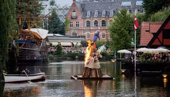 Ein kleines Boot fährt auf einem Kanal an einer brennenden Statue auf einem Holzfloß vorbei. In der Nähe versammeln sich Menschen mit Sonnenschirmen in einem Straßencafé. Im Hintergrund sind ein historisches Hochhaus und ein Großsegler zu sehen.