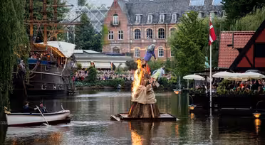 Ein kleines Boot fährt auf einem Kanal an einer brennenden Statue auf einem Holzfloß vorbei. In der Nähe versammeln sich Menschen mit Sonnenschirmen in einem Straßencafé. Im Hintergrund sind ein historisches Hochhaus und ein Großsegler zu sehen.