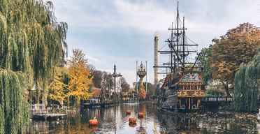 Ein malerischer Blick auf einen Freizeitpark mit einem alten Schiff, das an einem ruhigen See angedockt ist. Herbstfarben färben die Bäume am Rand, und skurrile Parktürme ragen vor einem blauen Himmel empor. Große orangefarbene Bojen schwimmen auf dem Wasser.