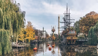 Ein malerischer Blick auf einen Themenpark mit einem Piratenschiff auf einem ruhigen See. Herbstbäume mit gelben und orangefarbenen Blättern säumen das Ufer. Dekorative Türme und Kürbisdekorationen sorgen für festliche Stimmung unter einem strahlend blauen Himmel.