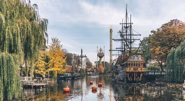 Ein malerischer Blick auf einen Themenpark mit einem Piratenschiff auf einem ruhigen See. Herbstbäume mit gelben und orangefarbenen Blättern säumen das Ufer. Dekorative Türme und Kürbisdekorationen sorgen für festliche Stimmung unter einem strahlend blauen Himmel.