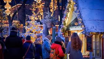 Eine festliche Marktszene im Freien mit Menschen, die an Ständen stöbern. Funkelnde Lichter schmücken Bäume und Gebäude und schaffen eine warme, einladende Atmosphäre. Ein schneebedeckter Stand trägt eine kleine Kiefer auf dem Dach, und Kunden haben sich um ihn versammelt.