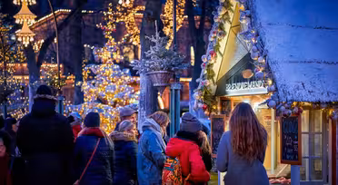Eine festliche Marktszene im Freien mit Menschen, die an Ständen stöbern. Funkelnde Lichter schmücken Bäume und Gebäude und schaffen eine warme, einladende Atmosphäre. Ein schneebedeckter Stand trägt eine kleine Kiefer auf dem Dach, und Kunden haben sich um ihn versammelt.